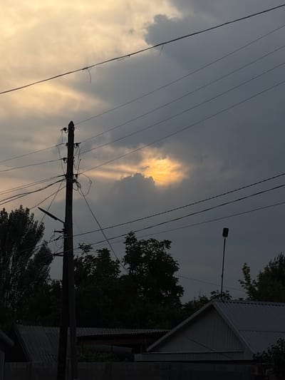 This image depicts a slightly overcast sky with dramatic cloud formations. A patch of bright, illuminated cloud is visible in the upper left quadrant, suggesting sunlight breaking through. Several power lines crisscross the frame, some appearing tangled. A utility pole with insulators and wiring stands prominently in the center, extending upwards. In the background, the corrugated metal roof of a building and dark green trees are visible on the right side. A street lamp is mounted on a pole in the upper right. The overall mood is somewhat somber due to the heavy clouds, but the illuminated cloud offers a touch of hope or drama. The scene appears to be set in a residential or semi-urban area, possibly in the late afternoon or early evening given the quality of light. There is no visible text in the image. The rotation of the image suggests it may have been captured vertically.