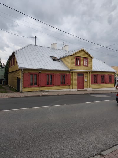 An exterior, eye-level, full shot captures a traditional, yellow wooden house with a silver, pitched roof in Zgierz, Poland. The house features red-shuttered windows and red double doors. To the left of the house, a dark green hedge and garbage bins are visible. The house is situated alongside a gray asphalt road, separated by a pedestrian sidewalk made of red and gray pavers. Overhead, a cloudy sky is crisscrossed by electrical wires.
