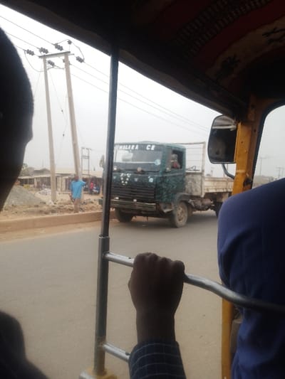 The image depicts a street scene in Lapai, Nigeria, viewed from inside a moving vehicle, likely a keke napep (tricycle taxi), given the visible internal structure and patterned ceiling.
In the immediate foreground, to the left, the dark-skinned scalp and upper portion of an individual's head are visible. To the right, a dark-skinned hand, wearing a blue and white checkered sleeve, firmly grips a horizontal chrome-plated metal bar, which is part of the vehicle's internal frame. Further right, the upper back and shoulder of another individual, dressed in a solid blue garment, are partially visible. The vehicle's yellow frame and a patterned fabric lining the ceiling are also apparent.
Through the vehicle's window, an asphalt road occupies the midground. A dark green and white mottled flatbed truck, appearing aged, is approaching from the opposite direction. The truck bears the inscription "THALARA CE" above its windshield, and a person is visible in the driver's seat.
In the background, along the right side of the road, a dirt shoulder with construction debris and dirt piles is present. A male individual wearing a light blue shirt stands facing left on this shoulder. Further back, multiple utility poles with overhead wires extend into an overcast, light gray sky. Distant, indistinct structures and buildings suggest a populated urban or semi-urban environment.