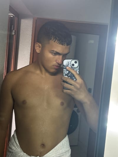 A young man is taking a selfie in a bathroom mirror. He is shirtless and wearing a white towel around his waist. His skin appears to be damp. He is holding an iPhone in his left hand with a black and white case. The background shows a bathroom with a white wall and a possible washing machine. The location is unknown, as is if the image was taken in the city of Septemvritsi, Bulgaria.