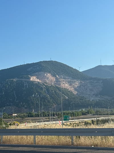 The image shows a mountain adjacent to a highway near Kırkağaç, Turkey. The mountain has visible rock formations and sparse vegetation. Wind turbines are situated on the higher elevation. On the right of the mountain, there is a highway with signs. A guardrail separates the road from an adjacent field. The sky above is clear and blue.