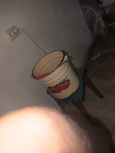 In Perliavka, Ukraine, a close-up, low-angle shot captures a metal bucket with a strawberry design, positioned on a dark wooden stool. The bucket, cream-colored with a black rim and a red handle, is adorned with illustrations of red strawberries and white flowers. A teal cloth is draped beneath it on the stool. The background reveals a white wall with an electrical outlet and a black cord running towards a power strip. The floor is made of grey wood planks, and shadows are cast on the wall and floor. The scene is dimly lit, with a large, out-of-focus blur of light dominating the foreground, obscuring any potential human presence.