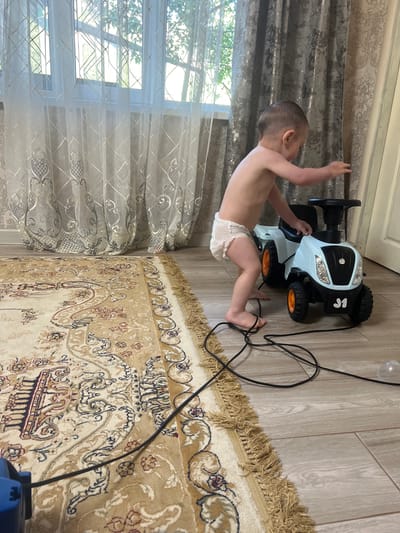 A young child, wearing a diaper, is captured mid-movement, appearing to be reaching out or about to move. The child is positioned next to a small, light blue toy car with black wheels and orange accents. A black cord, likely belonging to the toy car, snakes across the floor and onto a patterned rug. The floor is made of light-colored wood-look planks.
The background includes a window covered by sheer, patterned curtains that let in a soft light, suggesting it might be daytime. The curtains have a decorative, almost ornate design. To the left of the child and the toy car, there are thicker, textured curtains, possibly in a greyish tone with a metallic sheen. A part of a blue electrical device is visible in the bottom right corner. The overall setting appears to be indoors, likely a home environment. The lighting suggests natural light filtering through the window. The child's pose and the presence of the toy car hint at playtime or an attempt to interact with the toy. There is no visible text in the image.