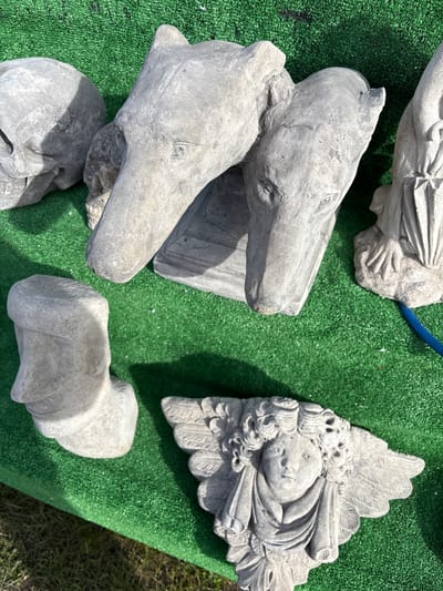The image shows several weathered, grey stone sculptures placed on a patch of bright green artificial turf. At the top-left is a partially visible draped female figure. Below it and more prominently displayed, are two horse head sculptures resting on what appears to be additional pieces of sculpture; all are made of stone. To the right is an angel wing sculpture. An additional piece of sculpture, partially obscured, is located at the bottom-left. The turf suggests an outdoor setting. No people are visible. The location in Chirk, United Kingdom, is based on additional non-image context.