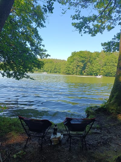 The image depicts a serene natural scene by a body of water, likely a lake or river. In the foreground, two portable camping chairs are set up on a muddy bank covered with grass and fallen leaves. Between the chairs, a thermal bottle and a plastic bag containing food items are visible. The water surface is rippled, reflecting the clear blue sky and the lush green trees that line the opposite shore. A dense forest forms a backdrop to the water, with the silhouettes of trees extending across the horizon. Two small boats are seen on the water in the mid-ground, with one actively moving and creating a white wake. The overall impression is one of a tranquil outdoor setting, suitable for relaxation or a picnic. Given the context, the location is identified as the vicinity of Olsztyn, Poland.