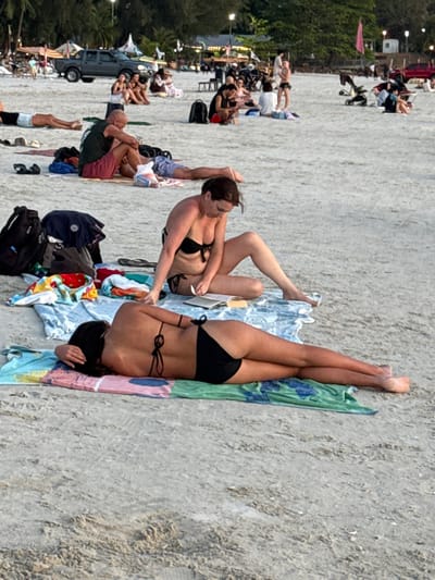This image depicts a crowded beach scene at Cenang Beach, Malaysia, likely during late afternoon or early evening.

In the foreground, two adult females are present. One female lies prone on a multicolored striped towel, facing away from the camera, wearing a black two-piece bikini. Her dark hair is visible. Immediately behind her, a second female sits cross-legged on a light blue towel, wearing a black two-piece bikini. She is actively reading an open book held in her hands. Adjacent to the sitting female are a black backpack, a pair of black flip-flops, and various folded garments on the sand.

In the midground, numerous other individuals are scattered across the sandy beach. A male figure wearing a dark green tank top and red shorts is seated on the sand, looking downwards. Another individual is seen lying on their back further in the midground. Personal belongings like bags and towels are interspersed among the groups.

The background shows more people relaxing on the expansive sandy area. A dark grey pickup truck is parked on the sand to the left. Further back, several structures, possibly beachside establishments, are visible, marked by illuminated lights. A row of green trees and foliage lines the far edge of the beach. Overhead streetlights are also discernable. A baby stroller is visible towards the right in the background. The overall lighting suggests ambient daylight transitioning towards dusk.