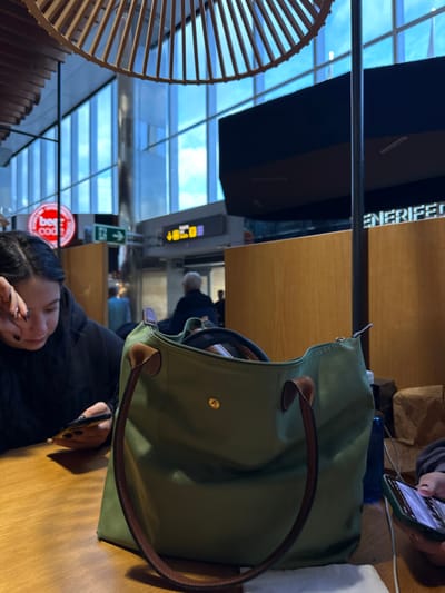 A young woman with long dark hair is sitting at a wooden table in a large, modern, brightly lit space. She is wearing a black hooded sweatshirt and is looking down at a smartphone in her hands. She is sitting on the right side of the table. A large, green tote bag is sitting on the left side of the table. The bag is made of nylon and has leather straps and handles. In the background, there are large windows that look out onto a bright, sunny day. There are several other people walking around in the space. The space looks like a modern airport terminal, likely in the Tenerife South Airport, located in the city of Granadilla de Abona, Spain, at the coordinates 28.048 latitude, -16.578 longitude. The woman is likely waiting for a flight.