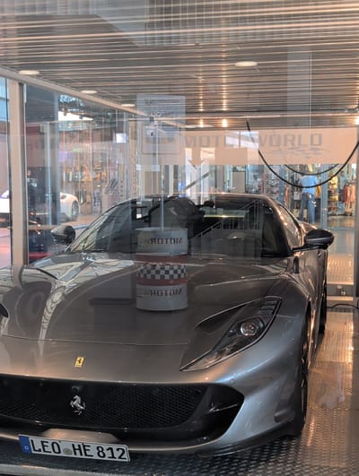 This is a medium shot of a gray Ferrari parked behind a glass window, possibly in a showroom or museum. The car is the primary subject, and its sleek design and metallic finish are highlighted by the lighting. The background shows a reflection of the interior, with displays of merchandise and possibly other cars.

Visible text includes "MOTORWORLD" printed vertically on a sign behind the car, and a license plate on the rear of the Ferrari that reads "LEO HE 812 D". There are also some checkered flag patterns visible on cylindrical objects in the background, likely part of the "MOTORWORLD" branding. The lighting appears to be artificial, suggesting an indoor environment. The car is stationary, and no people are clearly visible or actively engaged in any activity. The overall impression is one of a display of a high-performance vehicle. The location context is Böblingen, Germany.