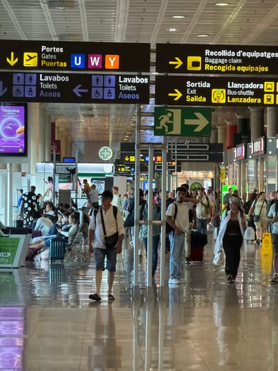 The image depicts an interior scene at Josep Tarradellas Barcelona-El Prat Airport (BCN) in El Prat de Llobregat, Spain. The airport environment is characterized by its modern architecture, with a high, beamed ceiling and bright, reflective floors. Overhead, a series of directional signs are prominently displayed, indicating various airport services and gates. These signs are bilingual, featuring Catalan and English text, and are accompanied by clear pictograms.
One sign, labeled "Portes Gates Puertas," directs travelers to gates U, W, and Y. Another indicates "Lavabos Toilets Aseos," with symbols for general, accessible, and family restrooms. Further signs point towards "Recollida d'equipatges Baggage claim Recogida equipajes" and "Sortida Exit Salida" with an arrow for "Llançadora Bus-shuttle Lanzadera" for terminal transfers. A green exit sign with a running figure and an arrow is also visible.
In the foreground, several people are walking and sitting. A man in the right foreground is walking away from the camera, wearing a white shirt, denim shorts, and dark sandals. A woman in the mid-right is walking towards the camera, wearing a white shawl over dark clothing. Other individuals are seated on airport chairs, some looking at their phones, while others are standing and conversing. A man in the mid-left is sitting with luggage. The lighting is bright, with fluorescent lights on the ceiling and a luminous advertisement screen on the left displaying "Shopping is in the air." The overall atmosphere is busy, typical of an active airport terminal.