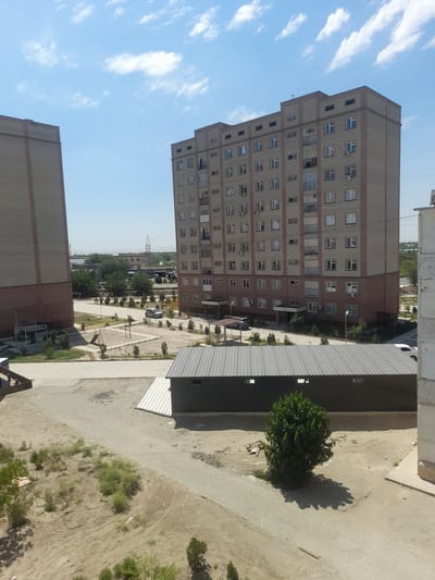 The image depicts a residential area in the city of Urgench, Uzbekistan. Two tall apartment buildings, constructed from light brown brick with darker brown trim, dominate the scene. The building on the right is fuller and has multiple floors with visible windows, balconies, and air conditioning units. The building on the left is only partially visible, appearing more slender. In the foreground, a low-lying, dark-colored building with a corrugated metal roof and visible lights sits on a sandy, unpaved area. A solitary green tree with a distinct shadow is located to the right of this structure. Scattered around the area are patches of dry vegetation and some debris. A playground structure with a slide is visible in the middle ground, near a paved pathway. Several cars are parked or driving on the paved surfaces. The sky is a clear blue with scattered white clouds. The overall impression is that of a moderately developed urban environment under a bright, sunny sky.