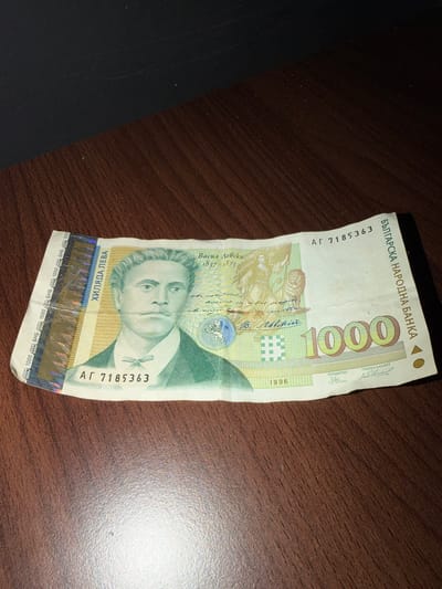 The image shows a single 1000 Bulgarian Lev banknote, dated 1996, lying on a wooden surface. The banknote features an image of Vasil Levski, a Bulgarian revolutionary, and the text "БЪЛГАРСКА НАРОДНА БАНКА" (Bulgarian National Bank). The serial number on the banknote is АГ 7185363. The bill is slightly crumpled and worn. There is no indication that the photograph was taken in Radomir, Bulgaria.