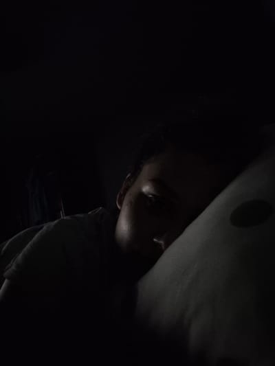 An individual is depicted resting in a dimly lit interior environment within Iskrets, Bulgaria. The subject lies on their left side, with the left portion of their face, encompassing an eye and part of the nose, partially illuminated. The remainder of the face and body are cast in deep shadow. A light-colored pillow featuring a prominent dark, circular marking is situated to the right of the subject's head. The surrounding background is uniformly dark, precluding the identification of specific environmental details. The individual appears to be in a state of repose.
