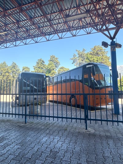 A view of two buses parked behind a blue metal fence on a paved lot. The upper bus is primarily orange and the lower bus is gray. Trees are behind the buses. A metal frame structure is visible to the left. The location is Polje, Montenegro.