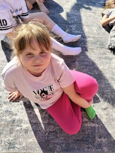 In this overhead shot, a young girl with blonde hair sits cross-legged on a gray patterned rug, bathed in bright sunlight. She's wearing a light pink t-shirt with a graphic of the word "VOGUE" and sunglasses-wearing figures, along with bright pink leggings and a green sock on her left foot. Her hands are resting on her knees, and she looks directly at the camera with a slight smile, her eyes a striking blue-green. The shadow of what appears to be a tree or structure falls diagonally across the scene, suggesting it's daytime and outdoors.

In the background, other children are also visible, suggesting a gathering or playtime. To the left, a child wears a white t-shirt with black stripes on the sleeves and the number "10" printed on the front, along with white socks. Further back, another child's legs are visible, also wearing white socks. To the right, a child in a patterned t-shirt and denim shorts lies on the rug. The presence of multiple children, casual attire, and the sunny setting indicate a relaxed, informal event, likely outdoors on a pleasant day. The scene is set in Staryi Aidar, Ukraine.