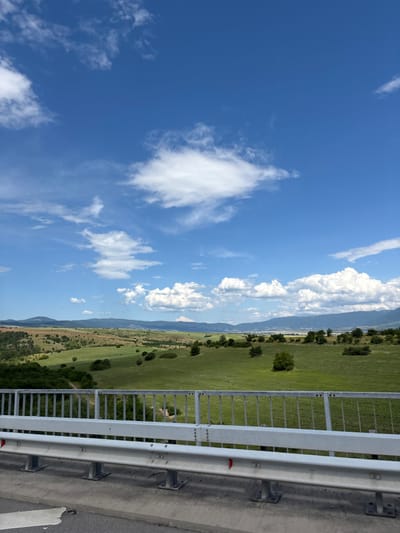 The image depicts a scenic landscape from a slightly elevated perspective, likely from a bridge or overlook. In the foreground, a sturdy white metal guardrail with vertical bars and a horizontal beam runs across the bottom third of the frame, suggesting a roadway or pathway. Beyond the guardrail, a vast expanse of rolling green hills unfolds, dotted with scattered trees and shrubs. The hills are bathed in sunlight, highlighting the vibrant green of the fields and the subtle variations in the terrain. In the distance, a range of mountains forms a hazy backdrop against a bright blue sky filled with fluffy white clouds. The scene conveys a sense of natural beauty and openness, characteristic of a rural or semi-rural environment. No people or specific landmarks are visible to pinpoint an exact location within Blatino, Bulgaria. The image captures a serene, natural vista on a clear day.