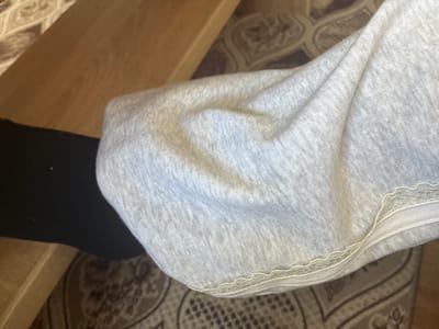 The image shows a close-up view of a person's legs and clothing, as well as a wooden surface and a carpet. The person is wearing a black sock on their left leg and a light gray garment, possibly a pair of sweatpants or a skirt, on their right leg. The garment has a lace trim along the bottom edge. The wooden surface appears to be part of a table or chair. The carpet has a patterned design in shades of brown and beige. The scene is indoors and appears to be a domestic setting. There are no identifying landmarks or contextual clues to suggest a specific location in Radievo, Bulgaria.