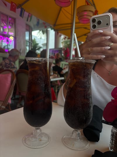In this image, two tall glasses filled with dark liquid and ice, likely a cola beverage, are positioned on a white table. White straws are visible in both glasses. The glasses have a unique, bulbous base that tapers towards the rim. A person's hands are visible at the top of the frame, holding a smartphone, suggesting they are taking a selfie. One hand, adorned with rings and manicured nails, is near the phone.
In the background, out of focus, people are seated at tables, and a yellow umbrella shades the area, indicating an outdoor setting, possibly a restaurant or cafe. Small flags are visible in the lower left corner, including what appears to be the Turkish flag, suggesting a diverse or international clientele. The lighting suggests it is daytime, possibly afternoon. The overall mood appears casual and relaxed. The image appears to be taken in Miami Beach, Florida, USA. There is some neon signage visible in the background with glowing purple and yellow letters, but the text is not entirely legible.