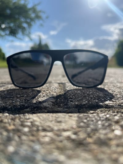 A low-angle, close-up shot of black sunglasses sitting on a textured, light-colored surface that appears to be asphalt or concrete. The sunglasses are the primary subject, with their dark lenses reflecting a bright, hazy sky with scattered clouds. The background is out of focus, showing hints of green foliage and trees on either side, and a bright sun flare in the upper right corner. The overall impression is an outdoor, possibly urban or suburban setting, bathed in bright sunlight.