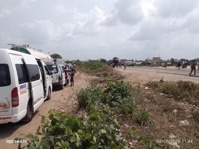 The image depicts a roadside scene in Ode, Nigeria, recorded at 11:44 on February 6, 2026, under a mostly cloudy sky. On the left, a queue of vehicles occupies a dirt shoulder, primarily consisting of white passenger vans, including one prominently displaying "ORIZU" and an emblem with its sliding door open. Behind the vans, a large white tanker truck is visible. Several individuals are standing or walking near these vehicles, some appearing to interact with them. The foreground features green leafy vegetation.
The right side of the image presents a wide, unpaved road or shoulder, transitioning into a darker paved section further right. Multiple pedestrians are scattered across this area, with one individual in a green outfit walking on the paved portion from right to left. In the mid-distance, a brown and yellow sign with "SAVE OIL" and additional text is visible adjacent to the road. The background contains various low-rise buildings and a tall communication mast. The ground on the right consists of dry earth, sparse vegetation, and scattered refuse.