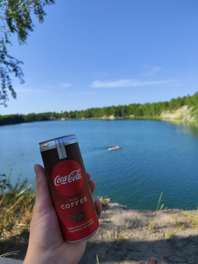 A close-up, slightly downward angled shot shows a person's left hand holding a can of Coca-Cola Plus Coffee. The can is predominantly dark brown with a red Coca-Cola logo and text indicating "PLUS COFFEE" and coffee bean imagery. The background features a bright blue sky, a serene, blue body of water, and a lush green treeline with some sandy cliffs. In the distance, two individuals are visible in the water, one of whom appears to be swimming. The scene evokes a sense of leisure and outdoor activity, possibly at a lake or quarry. The provided metadata does not specify a location in Korosten, Ukraine.