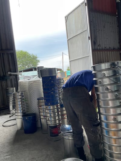 In Кенен Әзірбаев ауылы, Kazakhstan, a person wearing a blue t-shirt and grey work pants is bent over, appearing to interact with stacks of large metal and plastic buckets. The scene is indoors, within what seems to be a storage or workshop area, with a large corrugated metal door partially open, revealing a cloudy sky and trees outside. Several stacks of shiny metal buckets are prominent, along with some blue plastic buckets and large white plastic containers. The floor is concrete, and there is evidence of industrial or construction materials. The overall impression is one of a work in progress or a storage facility.