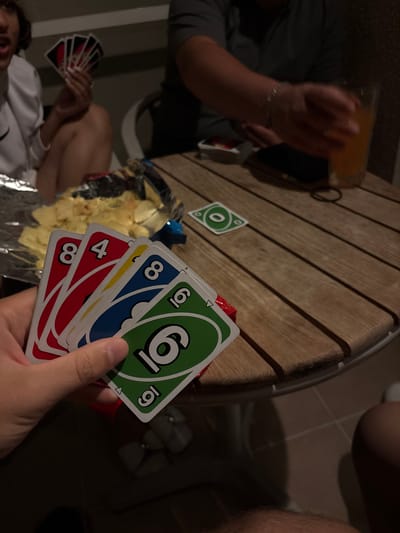 This dimly lit image captures a casual social gathering centered around a game of Uno, likely taking place during the evening or night in Selçuk, Turkey. In the foreground, a person's hand holds a fan of Uno cards, clearly showing a red 8, a red 4, a yellow card, a blue 8, and a green 6. A small triangular symbol is also visible on the green 6 card.
On the round, slatted wooden table, a green Uno card with a '0' and the "UNO" logo is face up, indicating it's the card in play. Nearby, a bag of potato chips rests on the table. To the upper left, another participant's arm, adorned with a silver bracelet, holds a glass containing an amber-colored beverage. In the lower left, the hand of a third player is visible, holding their own Uno cards, predominantly red, while wearing a white top.
The setting appears to be a relaxed indoor or outdoor patio area, evidenced by the wooden table and a light-colored tile floor. The artificial lighting casts shadows, contributing to the intimate, evening atmosphere of the gathering. The scene depicts a moment of enjoyment and social interaction centered around the card game.