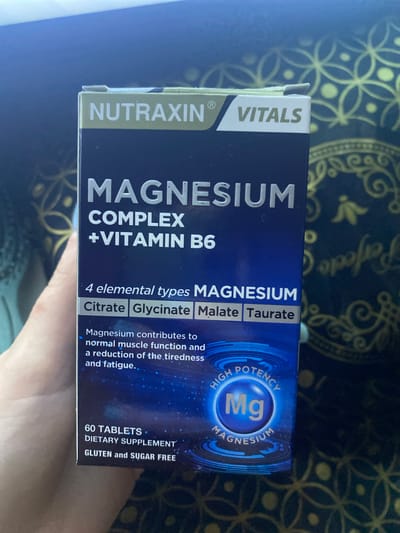 A box of Nutraxin® Vitals Magnesium Complex + Vitamin B6 dietary supplements is prominently displayed, held by a person's hand which is partially visible at the bottom and left of the frame. The box is oriented sideways, with its front panel facing the viewer. The packaging is primarily dark blue with white and light blue text and graphics.

Visible text on the box includes the brand "NUTRAXIN® VITALS" at the top left. The main product title reads "MAGNESIUM COMPLEX + VITAMIN B6". Further details specify "4 elemental types MAGNESIUM: Citrate, Glycinate, Malate, Taurate." A descriptive text states: "Magnesium contributes to normal muscle function and a reduction of the tiredness and fatigue.." At the bottom right, it indicates "60 TABLETS", "DIETARY SUPPLEMENT", and "GLUTEN and SUGAR FREE". A circular graphic on the right side features "HIGH POTENCY Mg MAGNESIUM".

The background behind the box is a dark surface, possibly fabric or a tabletop, adorned with a repeating gold or yellow circular and floral pattern. The lighting appears to be indoors.
