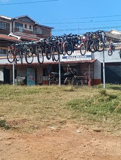 The scene depicts an exterior view in Ndenderu ward, Kenya, under a clear blue sky. In the foreground, an unpaved, reddish-brown dirt surface with scattered small stones transitions into a strip of dry and green grass. The midground features a large, horizontal metal rack, supported by white vertical poles, from which approximately 30-40 bicycles of various models and sizes are suspended. Behind this bicycle display is a single-story commercial building with a corrugated metal roof. A prominent blue and white sign on the building's facade reads "OPEN UNIFORMS RUAKA BRANCH" followed by phone numbers +254 706 841 896 and +254 721 864 881. To the left, a partially obscured sign indicates "AGGIES SALON & COSMETIC". Two dark-colored motorcycles are parked on the ground in front of the building, beneath the hanging bicycles. In the background, a multi-story building with a brown corrugated roof and multiple windows is visible. Overhead, several utility wires are strung across the frame. No individuals are present, and no specific actions or interactions are depicted.