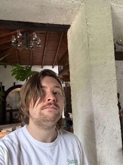The image is a selfie taken from a low angle. A person with long, light brown hair and a beard is in the foreground, wearing a white t-shirt. In the background is an interior scene with a rustic wooden ceiling and what appears to be a chandelier hanging from it. A large monstera plant is also visible. The background also contains hints of furniture and other decorative elements such as a mirror and framed artwork, but these details are not entirely clear. The location is likely inside a building, possibly a home, in Cañada de Madero, Mexico.