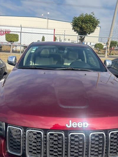 The image shows the front of a maroon Jeep parked outdoors. The Jeep's grill and "Jeep" emblem are visible. The windshield reflects the interior and the surrounding environment. A chain-link fence is in the background, along with a white warehouse-style building with "ES TLECUITL" partially visible. The location can be inferred to be Sanctorum, Mexico, based on the context provided. There are some trees and bushes visible behind the fence.