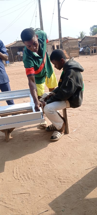 This image captures an outdoor scene in Ago-Abo, Nigeria, under direct sunlight, displaying two males engaged in an assembly task. In the foreground, a white, rectangular aluminum frame, resembling a window or door frame, rests on a rudimentary wooden table. The male on the left, standing, wears a green, yellow, and red striped t-shirt with yellow shorts; he is leaning over the frame, utilizing his right hand to manipulate a component. The male on the right is seated on a wooden stool, wearing a dark hooded sweatshirt and light grey sweatpants; he is intently focused downwards, appearing to work on a smaller object in his lap. The ground consists of reddish-brown, bare earth, marked with footprints and tire tracks. In the background, several informal wooden structures with corrugated roofs are visible, along with multiple indistinct individuals. Two wooden utility poles with overhead wires are prominent, and distant vegetation is visible against a clear sky.