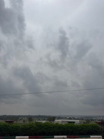 A wide-angle, eye-level outdoor shot shows a cloudy sky over Birnin Yero, Nigeria. The sky is filled with thick, gray clouds, suggesting an overcast day. A dark horizontal line, likely a wire, stretches across the middle of the frame. Below the sky, a green hedge forms a dense barrier in the foreground, with a red, white, and green striped border at its base. Beyond the hedge, a cityscape is visible, characterized by numerous buildings and trees, indicating an urban or semi-urban environment. A modern, light-colored building with a rectangular shape and prominent windows stands out among the other structures. The overall impression is one of a dull, cloudy day in a Nigerian city.