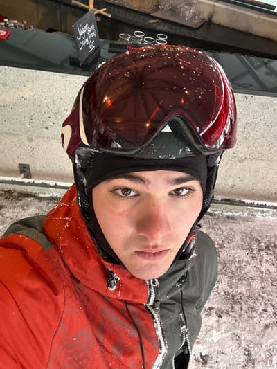 An image taken in Bad Hofgastein, Austria, captures a young male in winter ski attire, facing directly towards the camera. He wears a black balaclava, a red and black ski helmet with dark-tinted goggles pushed upwards, and a two-toned ski jacket, red-orange on his left side and dark grey on his right. Snow and ice particles are visible on the helmet, goggles, and jacket. His face shows exposed skin with light stubble on the chin and slight redness on the cheeks. The immediate foreground consists of snow and ice-covered ground with disturbed patches. In the background, a light grey textured wall is visible, above which a dark, flat surface or counter extends. On this counter, several pairs of clear glasses are arranged, and a small black chalkboard sign, suspended by twine, displays "Jäger bomb," "Flying mo*k*," and "6€" in white script.