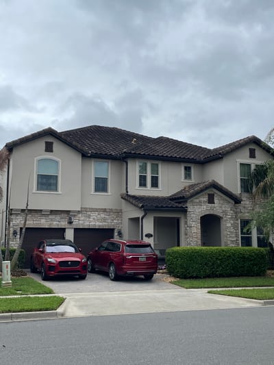The image shows a two-story house in Windermere, Florida, United States. The house features a combination of beige stucco and stone facade, with a dark brown tiled roof. There are multiple windows with white trim visible on both floors. A manicured green bush lines the front of the house near the entryway. Two red Jaguar vehicles are parked in the driveway. The sky is overcast. A sidewalk and road are adjacent to the property.