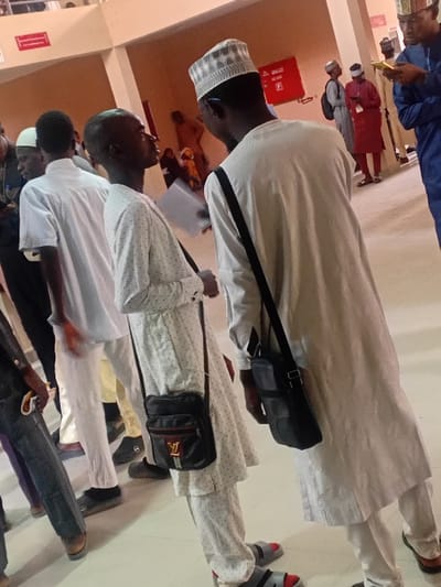 This image captures an indoor scene in the city of Kwazailewa, Nigeria, depicting a gathering of individuals within a modern public building. In the foreground, two men are central to the composition. The man on the left, with dark skin and short hair, wears a white tunic with small dark spots, white trousers, and grey-and-red sandals. A black messenger bag with a gold "LV" logo and a red-and-green stripe is slung over his left shoulder. He holds a white document in his left hand and looks upwards towards the man on his right. This second man, also with dark skin, is seen from a partial back view, wearing a plain white tunic, white trousers, and a white patterned kufi cap. A black messenger bag is slung over his left shoulder.
In the mid-ground and background, numerous other individuals are visible. Most wear traditional Nigerian attire, primarily white or light-colored tunics and trousers. One man in the mid-ground on the right wears a red tunic, while another on the far right wears a blue tunic and is engrossed with a yellow object, possibly a mobile phone. The environment features light-colored walls and flooring, suggesting a well-maintained hall or institutional setting. A red fire extinguisher is mounted on the wall in the mid-background, below a red sign with white text. The overall scene conveys a sense of congregation or passage within a public space.