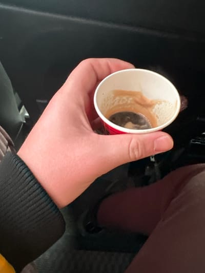 The image displays a light-skinned human hand holding a small, white paper cup with a red base, observed within the interior of a vehicle in the city of Ivanski, Bulgaria. The cup contains residual dark liquid, consistent with espresso or coffee, and light brown crema stains adhere to its inner walls. The subject's wrist is partially visible, encased by a dark, ribbed textile cuff. Below the hand and cup, a segment of dark brown fabric, likely part of trousers or a skirt, is discernible. The immediate background consists of dark, unidentifiable surfaces typical of an automobile's interior.