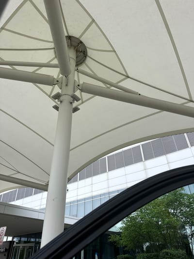 An upward angle shot shows the underside of a white tensile fabric structure supported by a central white pole and radiating white beams. The fabric is stretched taut and appears to be a canopy over an outdoor area. In the background, a modern building with a facade of glass windows and horizontal white louvers is visible. Trees with green foliage are reflected in the windows. A black car door frame or window trim curves into the foreground from the lower right. A red and white parking sign with a circle and diagonal line over a "P" is visible on the left side, along with the word "infant" on a sign below it, suggesting the location might be near a hospital or medical facility in Hoffman Estates, United States of America.