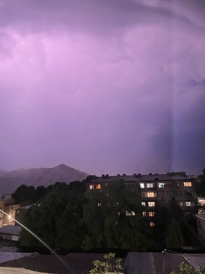 This media file depicts a nighttime scene in Gladzor, Armenia, characterized by a dramatic thunderstorm. The sky is filled with ominous purple clouds, illuminated by flashes of lightning. In the foreground, the edge of a building with a corrugated metal roof is visible, suggesting an urban or semi-urban environment. Adjacent to this roofline, lush green trees are silhouetted against the stormy sky.

The main subject of the image is a multi-story apartment building, its facade punctuated by numerous windows, many of which are lit from within, indicating residential activity. The building appears aged, with visible architectural details and what might be antennas on its roof. The presence of the building and the trees suggests a residential area, possibly with a view of the natural landscape.

In the distance, a mountain range is faintly visible through the haze and storm clouds, its contours softened by the low light and atmospheric conditions. The overall mood is dramatic and slightly unsettling, conveyed by the violent weather and the dark, nocturnal setting. The time of day is clearly nighttime, amplified by the artificial lights from the building's windows contrasting with the natural darkness. The weather is a significant element, with the thunderstorm creating a dynamic and visually striking atmosphere. No text is visible in the image.