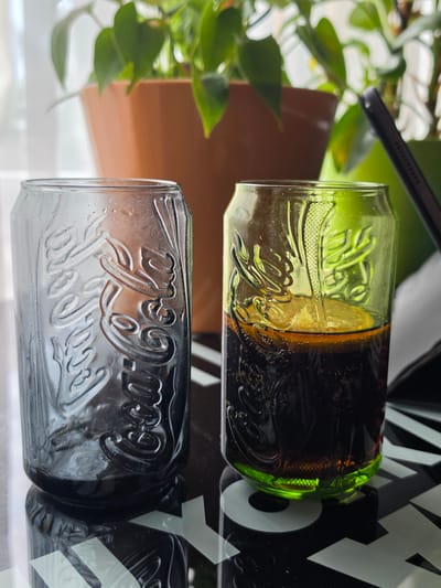 The image shows two Coca-Cola branded glasses. The top glass is green and contains a dark liquid, likely Coca-Cola, with a slice of lemon. The bottom glass is clear and appears to be empty, or nearly empty, with condensation visible on the exterior. Both glasses have the embossed Coca-Cola logo. The glasses are positioned horizontally, side-by-side, and reflect on a glossy surface below. In the background, out of focus, is a green plant with lush leaves and a brown object, possibly a planter. A dark tablet device is also partially visible in the upper left corner. The scene appears to be indoors, near a window, with natural light illuminating the subjects. The overall impression is one of refreshment and leisure. The text "Coca-Cola" is clearly visible on both glasses.