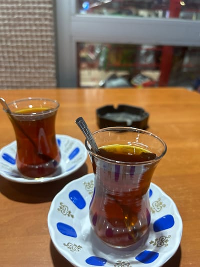 The image shows two traditional Turkish tea glasses filled with amber-colored tea, each resting on a small, decorative saucer. Both glasses contain a metal spoon. The saucers have a white base with blue oval shapes and a golden ornamental pattern. In the background, an ashtray is visible on a wooden table. Beyond the table, a window displays a blurred view of what appears to be a shop interior with shelves and products, possibly indicating the scene is set in a cafe or a similar establishment. The lighting suggests an indoor setting, possibly during the day given the light coming from the window. The overall scene evokes a sense of casual social gathering or relaxation. The location in Bayrampaşa, Turkey, contextually aligns with the cultural practice of drinking tea in such settings.