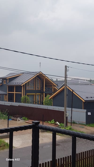 The image depicts a street scene in the Dolche Vita Village, Russia, on a cloudy day. In the foreground, a dark metal railing is visible, with water droplets on it, suggesting it is raining or has recently rained. Beyond the railing, a brown wooden fence separates the immediate area from a yard. The yard appears wet and shows signs of recent rain, with puddles on a paved surface. There are also stacks of lumber and some green vegetation.
In the midground, several modern houses with dark gray roofs and black wooden siding are prominent. These houses feature distinctive light yellow trim around the windows, balconies, and roof edges. One house, in particular, has a large, angled roof with expansive windows and a covered balcony. A telecommunications tower is visible atop one of the houses. A utility pole with wires stands to the right of the houses.
In the background, more rooftops of houses are visible, suggesting a residential development. A street sign with the inscription "8A" is also present near the utility pole. The overall atmosphere is overcast and slightly damp, consistent with rainy weather. No people are visible in the image.