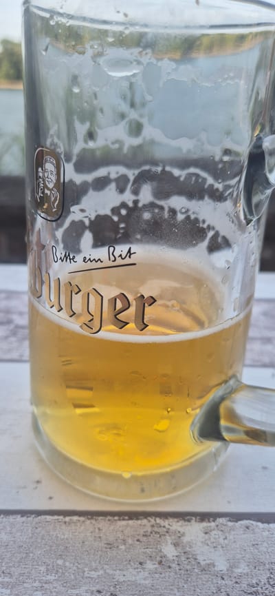 A close-up, vertical shot focuses on a nearly empty glass beer mug sitting on a white wooden table. The mug contains a small amount of pale yellow beer with some foam clinging to the sides. The glass is wet with condensation. A logo depicting a man's face in a gold circle is visible on the left side of the mug, and below it, the German text "Bitte ein Bit" is handwritten. Below that, the word "Burger" is printed in a gothic font. The background is blurred, showing a body of water and some trees, suggesting an outdoor setting. Given the context of "Burger" and "Bitte ein Bit," the image may be associated with a Burger restaurant chain or a specific location in Mainz, Germany, known for its beer culture.