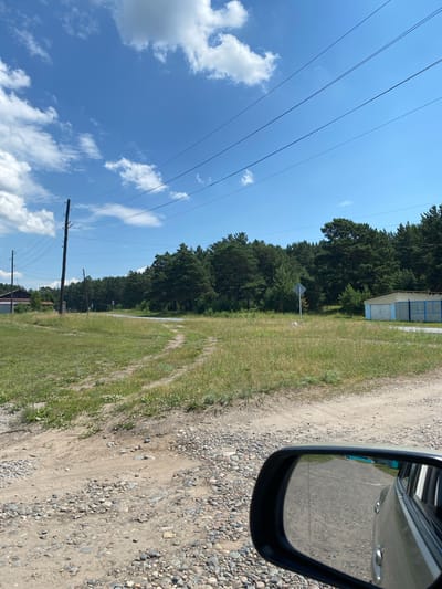 The image depicts a rural landscape under a bright, cloudy sky. In the foreground, a gravel and dirt road is visible, with tire tracks indicating recent vehicle passage. To the left, a grassy field with scattered wildflowers stretches towards a cluster of wooden buildings and power lines. A dirt path, also with tire tracks, branches off into the field. In the middle ground, a paved road curves through a field and disappears into a dense forest of pine trees. A single, diamond-shaped blue and white sign is posted in the field near the road. To the right, a light-colored building with a blue trim and a blue metal fence stands at the edge of the forest. The reflection in the car's side mirror shows the continuation of the gravel road. This scene is consistent with the outskirts of the village of Znamenka, Russia, characterized by its semi-rural setting with residential structures, unpaved roads, and surrounding natural vegetation.