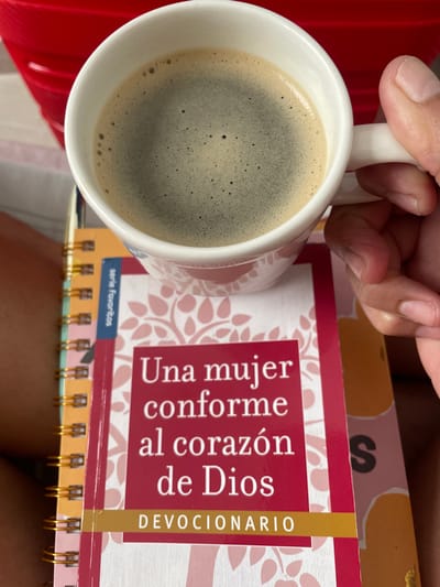 A close-up, top-down shot shows a person's hand holding a white mug of dark coffee. The mug is positioned to the left of a book titled "Una mujer conforme al corazón de Dios" (A Woman After God's Own Heart), which appears to be a devotional. The book sits atop a spiral-bound notebook. A red object is partially visible to the lower left of the mug. The environment and location are not discernable from the image, so the location cannot be identified as Rionegro, Colombia.