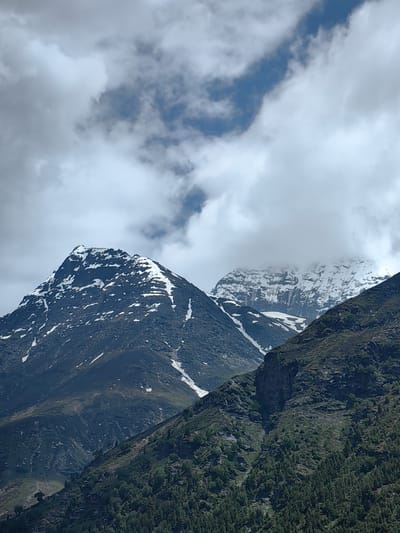 The image depicts a mountainous landscape with snow-capped peaks under a cloudy sky. In the foreground, a steep, rocky mountainside is covered with dense green vegetation, primarily trees, with patches of exposed rock. The midground features two prominent, dark-colored mountain peaks. The left peak is sharply pointed and has significant patches of snow scattered across its dark, rocky surface. The right peak is partially obscured by clouds but also shows snow cover, with a rugged, textured appearance. The background consists of a sky filled with a mix of bright white clouds and patches of blue sky. The lighting suggests it is daytime, with the sun likely behind the clouds, creating a diffused illumination. The scene is devoid of any people or man-made structures, presenting a purely natural environment. The location is identified as Burwa, India, suggesting this is a view of the Himalayan region.