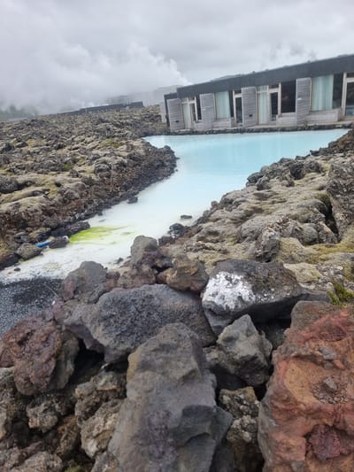 The image shows a view of the Blue Lagoon in Grindavík, Iceland. The scene features a body of milky-blue geothermal water bordered by rugged, moss-covered lava rocks. In the background, a modern, wooden structure with multiple windows is visible, suggesting a resort or spa facility. The sky is overcast with heavy clouds, indicating a cool or possibly misty day. The rocky foreground is detailed with various textures and colors, ranging from dark gray to reddish-brown volcanic rock, with patches of green moss. The water appears calm, with some lighter green algae or mineral deposits visible near the edge. The image is oriented sideways. No people or specific activities are discernible, but the setting evokes a sense of natural beauty and relaxation associated with geothermal spas. There is no visible text in the image.
