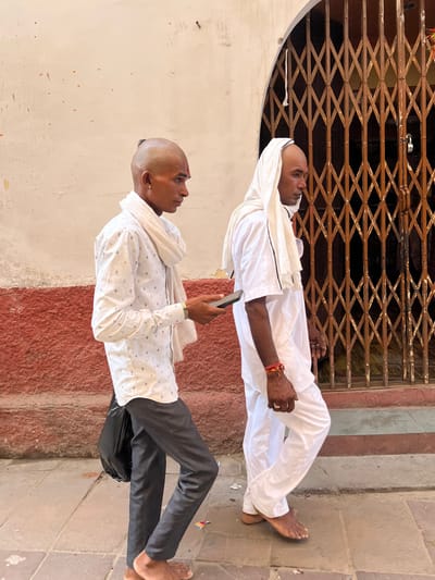 Two men are walking past a building with a large, intricate metal gate in Pushkar, India. The man on the left is wearing a white patterned shirt, dark pants, and a white scarf around his neck. He has a shaved head with a small tuft of hair at the back and is holding a mobile phone. He is also carrying a black bag. The man on the right is wearing a white outfit, including loose-fitting pants and a short-sleeved shirt, and has a white cloth draped over his head. Both men are barefoot. The building behind them has a textured reddish-brown base and a cream-colored upper section. The metal gate is ornately designed with diamond-shaped openings. The ground is paved with stone tiles.