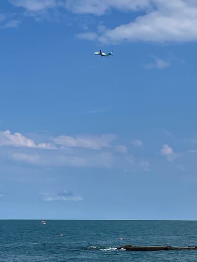 An airplane flies across a bright blue sky with scattered white clouds over a deep blue ocean. The airplane, a white passenger jet with blue accents, is in the upper portion of the frame. Below the sky is the ocean, which appears choppy with small waves. A small boat is visible on the water in the distance, as well as a red buoy and some submerged debris. The horizon line is clearly defined, separating the ocean from the sky. The scene is tranquil and open, with no indication of human presence in the immediate foreground. The location is described as Khostinskiy, Russia, a city on the Black Sea coast.