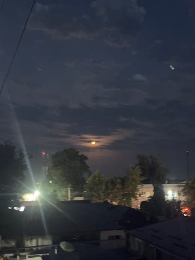 The image depicts a nocturnal urban scene, likely in Yangi'iul, Uzbekistan. The sky is dark blue with scattered clouds, and the moon is visible, partially obscured by the clouds and appearing with a yellowish-orange hue, suggesting it's either setting or rising through atmospheric haze.

The foreground is dominated by a dark, angled structure on the right, possibly a building or a wall, which obscures much of the view. To the left of this structure, a bright, artificial light source casts a strong beam, creating lens flare and illuminating the surrounding foliage. This light source appears to be a streetlamp or a building light.

Further into the scene, there are trees with green foliage. In the background, several buildings are visible, some with illuminated windows, indicating human habitation or activity. A traffic light with a red illuminated bulb is also visible. There are power lines in the lower part of the image. The overall impression is that of a quiet night in a city or town, with a mix of natural and artificial light. The presence of buildings, streetlights, and traffic lights suggest an urban or suburban setting. The time of day is clearly night.