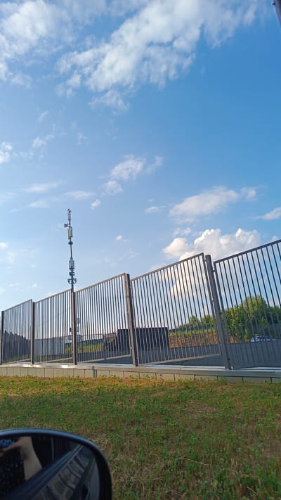 The image shows a cell tower behind a metal fence. The sky is blue with some white clouds. There is green grass in the foreground. The scene is likely from a public area or roadside in the city of Zvenigorod, Russia. No people are visible in the image, but there is a reflection of a hand in the side-view mirror of a vehicle, suggesting the photo was taken from a car. The fence appears to be part of a perimeter around a facility or property. The cell tower has multiple antennas and equipment attached to it.